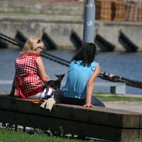 Two women and a small dog sit on a bench in Liepāja, enjoying a sunny day by the water, with moored boats and a calm river in the background, signifying a moment