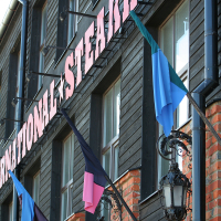 Facade of a steakhouse in Liepāja with bold signage, featuring black wood paneling and vibrant blue and pink flags under a clear sky, complemented by ornamental black street lamps.