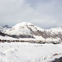 A serene winter landscape: a snow-covered village nestled among majestic mountains under a cloudy sky, with a gentle river cutting through the white expanse, known for its skiing and duty-free shopping in Livign