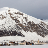 A tranquil alpine village nestles at the base of a majestic, snow-capped mountain in Livigno, with a slender church spire rising above cozy chalets under a cloudy winter sky.