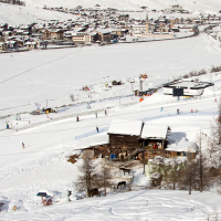 A snow-covered alpine village in the Italian Alps with ski slopes bustling with skiers, traditional buildings dotting the landscape, and a frozen river completing a picturesque winter sports destination.