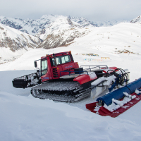 A snow groomer parked on a snowy mountain slope in Livigno, with a backdrop of rugged peaks, under a partly cloudy sky, ready to smooth the ski trails.