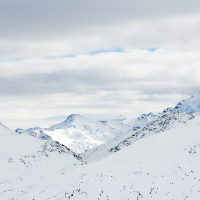 Snow-covered mountain peaks in Livigno, Italy, rise majestically under a cloudscape, with the sun gently highlighting the contours of the rugged terrain.