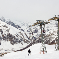 A skier descends a snowy slope with ski lifts overhead against a backdrop of majestic, snow-capped mountains under a cloudy sky, in Livigno, a renowned ski resort and vacation destination.