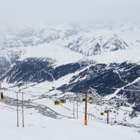 Yellow cable cars traverse above a snowy mountainous landscape in Livigno, with ski runs etched into the white slopes, amid a backdrop of overcast skies and distant alpine peaks.