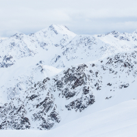 A majestic mountain range in Livigno, covered in snow under a cloudy sky, with the sun casting shadows and highlighting the rugged terrain's contours and peaks.