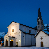A tranquil evening scene showing a snow-covered church with a pointed steeple, illuminated against the blue hues of dusk, in Livigno, Italy, with alpine mountains in the backdrop near the