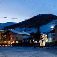 Twilight descends on a serene snow-covered village in Livigno with cozy alpine buildings, while distant ski slopes on the mountain await under a deepening blue sky, illuminated by the last vest