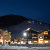 A tranquil alpine village at dusk, with warmly lit buildings against the backdrop of a snowy mountain and ski slopes illuminated for night skiing. Enjoy the unique charm of Livigno, renowned for its tax