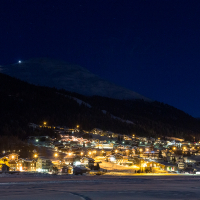 A tranquil mountain village in Livigno, Italy, aglow with warm lights under a twilight sky, with the serene presence of snow-capped peaks and a ski resort's illuminated trail in the distance