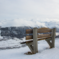 A solitary wooden bench overlooks a serene, snow-covered mountain landscape with the small village of Livigno nestled in the valley, offering a tranquil spot for contemplation amidst the beauty of a wintry