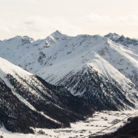 A serene winter landscape in Livigno showcases majestic snow-covered mountains with sunlit peaks, while a valley with sparse vegetation stretches below, hinting at a remote, tranquil location.