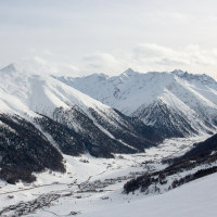 A snow-covered alpine landscape in Livigno, with a valley village, rolling hills dotted with evergreens, and majestic peaks under a cloudy sky.