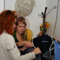 Two women examine clothing in a small, cluttered space. One woman, with curly red hair and glasses, holds a hanger with jeans. The other woman, with straight blonde hair and bangs, looks on. Behind them is a coat rack with hanging clothes, likely preparing for a Maiņas punkts preparty event.