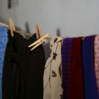 Several pieces of cloth, including a wooden clothespin-hung brown fabric, a floral-patterned beige scarf, and colorful garments in blue, red, and purple with a checked pattern, are drying on a clothesline indoors against a grey wall. The background includes a blurred framed Maiņas punkts sign.