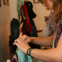 A woman with medium-length brown hair is looking closely at a green garment she is holding with both hands. She wears a black wristband and a colorful scarf. In the background, clothes on hangers and pegs are visible, hinting that this preparty shopping session might be happening at Maiņas punkts.