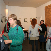 A room with white walls buzzes with preparty excitement as people engage in conversation and browse a clothing rack. A woman in the foreground, wearing a green sweater, holds a pair of pants. Framed artworks adorn the wall, and string lights hang above. To the right, a wooden door is partially open.