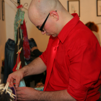 A bald man in a red shirt and tie, wearing black-rimmed glasses, is fixing or adjusting items clipped onto a line with colorful clothespins indoors. Framed pictures decorate the white wall behind him, adding an artistic flair to the preparty scene. Other people are present in the background.