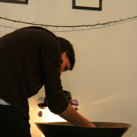 A person with dark hair, wearing a brown top, leans over a large, shallow basin on a dark table. They appear focused on something in the basin. Behind them, two framed abstract drawings are hung on a light-colored wall, and a string of lights decorates the space, setting the perfect preparty mood.