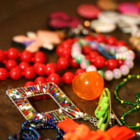 A close-up of assorted colorful jewelry pieces placed on a surface. In the foreground are beaded necklaces with red, green, and blue beads, a square pendant adorned with multicolored beads, and an orange, translucent bauble. Perfect for a preparty look, additional pieces blurred in the background include beads and various trinkets.