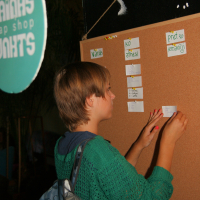 A person with short hair, wearing a green sweater and backpack, places a note on the corkboard at the maiņas punkts. The board has several small notes and the Latvian text "Variants, ko atnesi, Pret ko mainīji" written at the top. A round sign that is partially visible hangs on the left side.