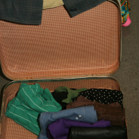 A partially packed open suitcase sits on the floor, containing an assortment of clothing. Visible items include a striped green shirt, a purple garment, a pair of jeans, a black pouch, and a red object. A gray checkered jacket is draped over the suitcase like it's ready for a preparty adventure, with colorful clothing visible in the background.