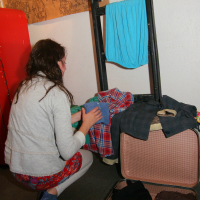 A woman with long brown hair, dressed in a light grey sweater and red skirt, is crouching down while packing clothes into an open suitcase. A ladder leans against the wall, draped with various garments, including a blue cloth and a checkered shirt. To the left stands a red wardrobe—readying for the preparty at her friend's maiņas punkts.