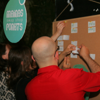 A group of people are pinning small paper cards to a large corkboard with pre-labeled sections at what seems to be a preparty. A bald man in a red shirt is prominently in the foreground. A teal sign above with white text reads "MARUAS maiņas punkts PUKHTS." Others in patterned clothing assist in organizing the cards.
