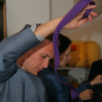 A man in a blue suit is holding a purple necktie with a checkered pattern above his head, seemingly preparing to tie it for the preparty. He is focused on the task. In the background, a woman is partially visible, also engaged in some activity. The setting appears to be indoors.