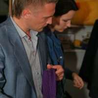 A man and a woman are standing indoors, both focused intently. The man, dressed in a light blue suit jacket and striped shirt, examines a purple tie with a grid pattern—a perfect choice for the preparty. The woman next to him, in a blue coat, is also looking downwards. Shelves with various items are visible in the background.