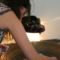 A person with long hair, wearing a sleeveless patterned top and a necklace, is bent over a large, round metallic tray filled with colorful pins and small items at the Maiņas punkts. In the background, a black cat mask with purple glasses resting on it is illuminated by a light source.