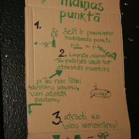A cardboard sign with green handwritten text in Latvian hangs on a wall. The title reads “Laipni lūgti Maiņas punktā” and includes four hand-drawn steps with illustrations: a package drop-off, additional item suggestions, finding new items, and registration information.