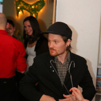 A person with a beard and mustache, wearing a black jacket and black cap, stands indoors looking slightly to the side. It appears to be a preparty at Maiņas punkts, with several people gathered behind them and a green decorative light shaped like a reindeer on the wall. A hand is holding a hanger near the person.