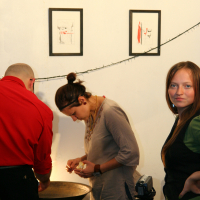 Three people in a room: one with short hair in a red shirt, another with a bun in a gray shirt peeling something at a table like it’s the preparty to an event at Maiņas punkts, and a third person with long hair in a black and green outfit looking at the camera. Two abstract paintings hang on the wall, and a string of lights runs between them. A tripod