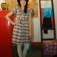 A woman with long dark hair wears a plaid dress, grey leggings, blue sandals, and a whimsical black hat with a purple bow. She stands in a room with a red refrigerator to her right, clothes hanging on a ladder behind her, and a decorative bust on the fridge. Smiling brightly, she seems ready for the preparty at Maiņas punkts.
