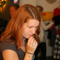 A young woman with long, red hair stands indoors, deep in thought with her hand near her mouth. She wears a gray t-shirt and colorful beaded jewelry. In the background, another person is visible at the maiņas punkts, sorting clothes on hangers. The room has soft lighting and a casual atmosphere.
