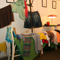 A cluttered room with various clothing items draped over furniture and hangers. On the left, a chair supports brown and green clothes. A green cushioned chair holds plaid, striped, and colorful shirts. A coatrack holds more clothing for the preparty prep. The background features wall art, string lights, and a table lamp.