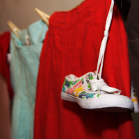 Various clothes are hanging on a clothesline secured with wooden clothespins. The items include a red dress, light blue shorts, a pair of white sneakers with colorful floral patterns, and a dark garment. Perfect for a preparty look, the sneakers are suspended by their laces, prominently featuring their vibrant design.