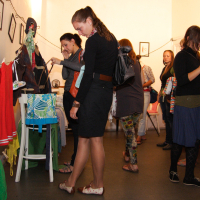 People browsing clothing and accessories in a small, indoor market or shop. The room is dimly lit with framed pictures on the walls. Clothes hang on a line and a rack. Individuals are engaged and examining items in this prepary Maiņas punkts. The atmosphere seems casual and friendly.