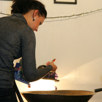 A woman with dark hair pulled back is standing at a table lighting a large candle in a shallow, wide bowl. She is wearing a long-sleeve grey top and dark pants, preparing for the preparty. The room is softly lit, with framed art on the wall and a string of fairy lights hanging in the background.