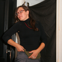 A woman wearing glasses and a black long-sleeved top pulls up her gray skirt. She stands in front of a black door and a black curtain, with a small blue light bulb illuminated above her. With long brown hair and a red beaded necklace, she exudes preparty vibes at this Maiņas punkts.