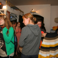 People shop at a crowded indoor market or Maiņas punkts. A person in a gray hoodie holds a striped skirt, while others browse nearby. Clothing hangs on racks, and various items are scattered throughout the space. The atmosphere appears lively and casual, as if preparing for a preparty.