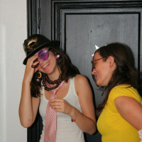 Two women are standing and smiling. The woman on the left is wearing a black hat, purple sunglasses, a white tank top, pink tie, and colorful pants. The woman on the right, sporting glasses with red horns and a yellow shirt, looks at her friend with amusement as they stand near a maiņas punkts against a black door.