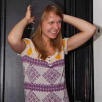 A young woman with light brown hair is smiling brightly while adjusting a gray beanie on her head in preparation for the preparty. She is wearing a white top with purple geometric patterns and yellow accents. Standing in front of a black door, she holds the beanie with both hands, her expression full of joyful amusement.