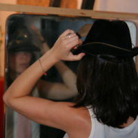 A person with shoulder-length dark hair is seen from behind, adjusting a black cowboy hat in front of a mirror. They are wearing a white sleeveless top. Their reflection shows a pink tie hanging loosely around their neck, preparing for the maiņas punkts preparty in a room with warm ambiance and artistic setup.