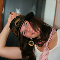 A woman with long brown hair smiles and tilts her head, holding the brim of a black hat at the Maiņas punkts preparty. She's wearing a white tank top, a pink patterned scarf, and a chunky necklace of large beads. An ornate decorative mask adorns her hat. A mirror and wooden frame appear in the blurred background.