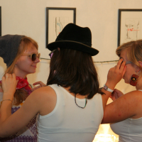 Three young women dressed casually in white tops are engaging in playful preparty activities. One wears a gray beanie and sunglasses, while a second ties a scarf around her neck. A third woman in a black hat helps another adjust a bandage on her arm. Framed art and string lights from Maiņas punkts adorn the wall behind them.
