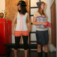 Two women are standing indoors in a room with eclectic decor, likely at a preparty. The woman on the left wears a black hat, white tie, white tank top, red patterned mini skirt, black leggings, and sandals. The woman on the right wears a grey hat, purple patterned top, grey skirt, boots, and holds a red cloth.