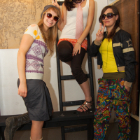Three young adults wearing colorful and eclectic outfits pose cheerfully in an indoor setting, perhaps at a preparty. Two stand on the ground, while one sits on a ladder. They all wear sunglasses and make peace signs, exuding a fun, playful vibe. A wall map and wooden furniture are visible in the background.
