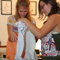 Two women are indoors, closely examining a white shirt. One woman with a colorful, sleeveless top and glasses is holding the shirt while the other woman in a patterned dress and a ponytail points to it. They seem focused and engaged, perhaps planning for their preparty. Several framed pictures hang on the wall in the background.